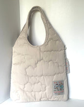 Load image into Gallery viewer, A light beige quilted puffer bag with cloud-like stitching, featuring a small colorful patch on the bottom right and a hanging tag, set against a plain white background.
