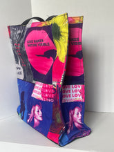 Load image into Gallery viewer, A tote bag made from printed magazine pages, featuring a collage of pink, blue, yellow, and black images with text like 'LOVE MAKES EVERYTHING WORTH WHILE' and 'LOVE IS VISIBLE', shown resting on a white surface.