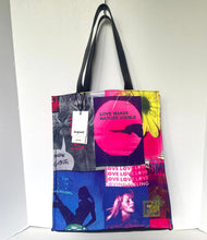 Load image into Gallery viewer, A tote bag with black handles and a vibrant collage print featuring pink, blue, yellow, and black graphic elements, hanging against a plain white wall.