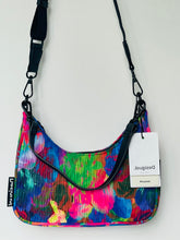 Load image into Gallery viewer, A colorful shoulder bag with a vibrant abstract floral pattern in pink, blue, green, and yellow, featuring black trim, a black adjustable shoulder strap, and a white hangtag.