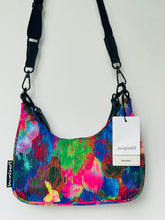 Load image into Gallery viewer, A small shoulder bag with a vibrant, multicolored abstract print featuring pink, blue, green, and yellow. It has black straps, black hardware, a white hangtag, and a black fabric label on the side.