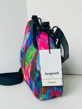 Load image into Gallery viewer, A colorful crossbody bag with a vibrant abstract paint stroke pattern in pink, blue, green, and purple, featuring black straps and a white hangtag with the brand name 'Desigual'.