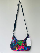 Load image into Gallery viewer, A colorful shoulder bag with a vibrant multicolor abstract print, featuring a black adjustable shoulder strap and a white product tag, hanging against a plain white wall.