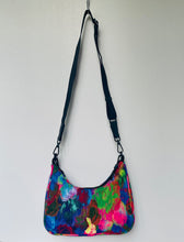 Load image into Gallery viewer, A crescent-shaped shoulder bag with a vibrant, multicolored abstract floral pattern in shades of blue, pink, green, and purple, featuring a black adjustable shoulder strap and black hardware, hanging against a plain white wall.