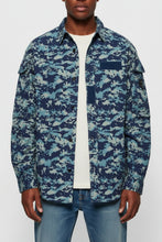 Load image into Gallery viewer, A man wearing a blue and light blue digital camouflage button-up shirt over a cream t-shirt and blue jeans, photographed against a light gray studio background.