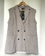 Load image into Gallery viewer, A sleeveless double-breasted blazer vest in a textured gray and beige tweed fabric, hanging on a wooden hanger against a white wall. The vest features notched lapels, a double-breasted design with black buttons, and front patch pockets.