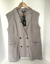 Load image into Gallery viewer, A sleeveless double-breasted blazer vest in a textured taupe-gray fabric, hanging on a wooden hanger against a white wall. The vest features notch lapels, a double-breasted button closure, and two front patch pockets.