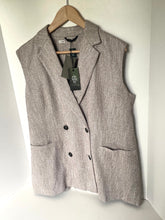 Load image into Gallery viewer, A sleeveless double-breasted blazer vest in a textured light gray-brown tweed fabric, hanging on a wooden hanger against a plain white wall. The vest features notch lapels, a double-breasted front with black buttons, and two front patch pockets.