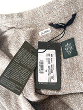 Load image into Gallery viewer, Close-up of a beige woven textile with attached retail tags, including a black hangtag, a white size tag, and a small green brand label reading ele ven ty.