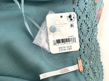 Load image into Gallery viewer, Close-up of a teal blue-green lace-trimmed garment with a white product tag and a small wooden brand tag reading 'L'Artisan'.