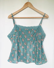 Load image into Gallery viewer, A light blue-green floral camisole top with thin straps and lace trim, hanging on a wooden hanger against a plain white wall.