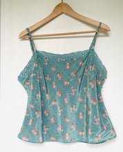 Load image into Gallery viewer, A light blue-green floral camisole top with thin straps and lace trim, hanging on a wooden hanger against a plain white wall.