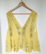 Load image into Gallery viewer, A pale yellow sleeveless top with a deep V-neckline, decorated with silver sequined geometric patterns, hanging on a wooden hanger against a white wall.