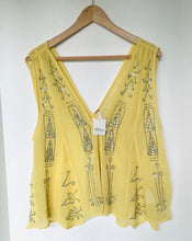 Load image into Gallery viewer, A pale yellow sleeveless vest with a deep V-neckline, decorated with silver rhinestone patterns, hanging on a wooden hanger against a white wall.