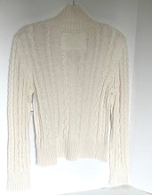 Load image into Gallery viewer, A cream-colored cable-knit turtleneck sweater hanging against a plain white wall.