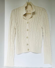 Load image into Gallery viewer, An off-white cable-knit cardigan sweater with a mock neck and button-up front, hanging on a wooden hanger against a plain white wall.