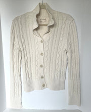 Load image into Gallery viewer, An off-white cable-knit cardigan sweater with a high stand collar and button-front closure, hanging on a wooden hanger against a plain white background.