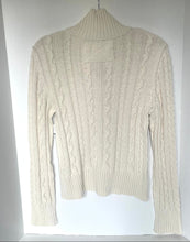 Load image into Gallery viewer, A cream-colored cable-knit turtleneck sweater hanging on a white hanger against a white background.