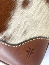 Load image into Gallery viewer, Close-up of a brown and white cowhide product with stitched leather trim and an embossed cross-like logo on a white background.