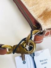 Load image into Gallery viewer, Close-up detail of a wristlet bag showing a brown leather strap, brass-tone hardware, a light tan faux-hair or fur trim, and a white product tag with instructions.