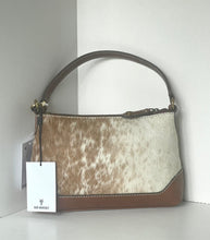 Load image into Gallery viewer, A handbag with a brown and white cowhide patterned body, brown leather trim and handle, and a white hangtag from 'Zip Yourself' on a white shelf against a white wall.