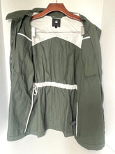 Load image into Gallery viewer, An olive green hooded jacket with white contrast panels and drawstring details, hanging on a wooden hanger against a plain white background.