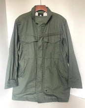 Load image into Gallery viewer, An olive green military-style utility jacket hanging on a wooden hanger against a plain white background.
