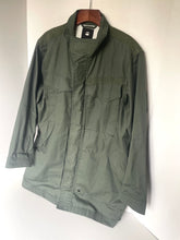Load image into Gallery viewer, An olive green military-style utility jacket hanging on a wooden hanger against a plain white wall.