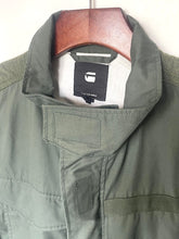 Load image into Gallery viewer, An olive green G-1000 jacket hanging on a wooden hanger against a plain off-white background. The brand label inside the collar reads G-1000.