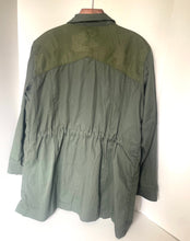 Load image into Gallery viewer, An olive green utility jacket with a contrasting darker green patterned yoke across the shoulders, hanging on a wooden hanger against a plain white wall.