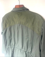 Load image into Gallery viewer, An olive green utility jacket with a textured woven yoke across the shoulders, hanging on a wooden hanger against a plain white wall.