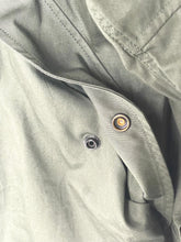 Load image into Gallery viewer, Close-up of an olive green fabric garment showing a buttoned pocket with a gold-toned snap and a black snap closure.