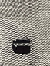Load image into Gallery viewer, A small black enamel pin shaped like a stylized letter 'G' on a textured gray woven fabric background.