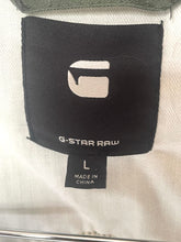 Load image into Gallery viewer, Close-up of a garment's interior label showing a black G-Star Raw brand tag with a white logo and size L, attached to a light beige textured fabric.