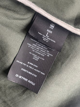 Load image into Gallery viewer, Close-up of a garment interior showing a black sewn-in size tag for G-Star Raw, labeled size L, attached to olive green fabric.