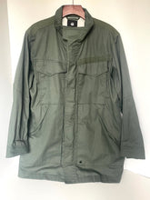 Load image into Gallery viewer, An olive green military-style utility jacket hanging on a wooden hanger against a plain white wall. The jacket features a high collar, a buttoned front closure, two chest flap pockets, and two large lower hand-warmer pockets.
