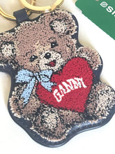 Load image into Gallery viewer, A teddy bear-shaped keychain with a textured, sequined appearance. The bear is brown and beige with a light blue bow and holds a red heart that says 'GANDY' in white.