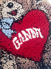 Load image into Gallery viewer, Close-up of a textured knit or woven fabric featuring a large red heart with the word "GANDY" in white, plus a light blue bow and a beige-and-black animal face motif.