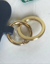 Load image into Gallery viewer, A close-up of a gold-tone metal key ring set attached to a dark navy blue leather strap, with a small clear plastic tag still connected. A partial green and white Simons tag is visible at the top edge.