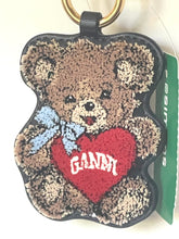 Load image into Gallery viewer, A teddy bear-shaped embroidered keychain or bag charm with a gold key ring, featuring a brown teddy bear holding a red heart with the word 'GANNY' in white, a light blue bow, and a black stitched border. A green hangtag is attached on the right.