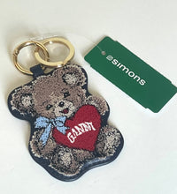 Load image into Gallery viewer, A Simons brand teddy bear keychain with a red heart that says 'GANNY' in white lettering, a blue bow, and gold key rings, shown next to its green Simons tag on a white background.
