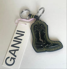 Load image into Gallery viewer, A black faux-leather cowboy boot keychain with yellow contrast stitching and a silver split ring, shown next to a white GANNI brand tag on a light off-white background.