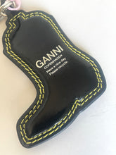 Load image into Gallery viewer, A black leather boot-shaped keychain with yellow contrast stitching and white printed text reading 'GANNI COPENHAGEN Have a nice day Please recycle.' A metal key ring is attached at the top, shown on a plain light background.