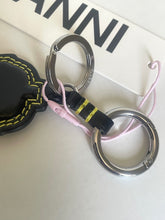 Load image into Gallery viewer, A close-up of a keychain with two silver metal key rings, a black fabric loop with yellow stripes, a light pink cord, and part of a black leather tag with yellow stitching. A white box with the brand name 'MANNI' is visible in the background.