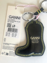 Load image into Gallery viewer, A black leather boot-shaped keychain with yellow contrast stitching and the brand name GANNI, shown next to its white paper tag on a light background.