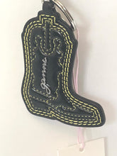 Load image into Gallery viewer, A black faux-leather cowboy boot keychain with yellow contrast stitching, a silver metal ring, and a pink hangtag on a white background.