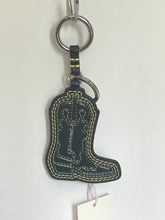 Load image into Gallery viewer, A black faux-leather cowboy boot keychain with light yellow contrast stitching and a silver metal key ring, shown against a plain off-white background with a paper tag attached.