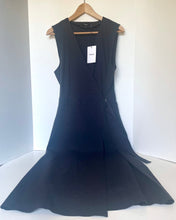 Load image into Gallery viewer, A sleeveless black wrap-style dress with a V-neckline and flared skirt, hanging on a wooden hanger against a plain white background. A white brand tag is attached near the neckline.