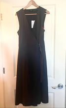 Load image into Gallery viewer, A sleeveless black wrap-style dress hanging on a wooden hanger in front of a white paneled door.