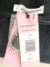 Load image into Gallery viewer, Pink retail tags and packaging for a dress by Avenue Spilles, including a white barcode label, a pink round tag reading "Dresses," and a clear plastic sleeve with decorative metal embellishments, placed on a black textured surface.